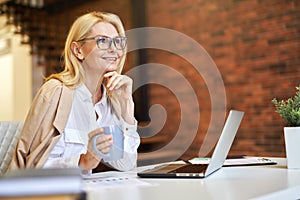 Cheerful mature businesswoman in glasses smiling away, using laptop and drinking coffee while working in the office