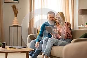 Cheerful Mature Couple Using Digital Tablet Relaxing Sitting At Home