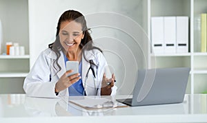 Cheerful mature lady doctor sitting in her cabinet, using phone