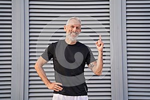 Cheerful mature man smiling at camera, raising hand and pointing index finger up while posing outdoors over gray