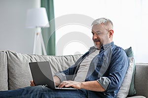 Cheerful mature man using laptop while resting on sofa