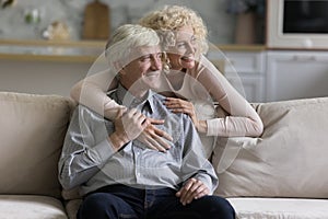 Cheerful mature wife hugging happy senior husband sitting on couch