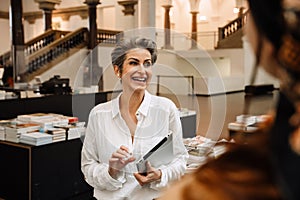 Cheerful mature woman art gallery manager holding tablet while standing in museum shop