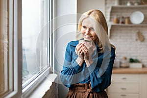 Cheerful mature woman enjoying morning coffee at home, standing near window at cozy kitchen, holding mug and smiling