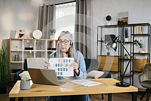 Cheerful mature woman presenting financial report to colleagues through video call on laptop.