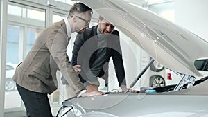 Cheerful men sales manager and client watching car engine under motor hood indoors