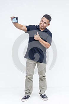 A cheerful middle-aged Asian man in casual attire takes a selfie and gives a thumbs up to the camera on a white background