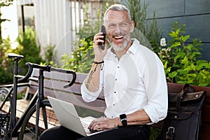 Cheerful middle aged businessman smiling, talking on the phone, using his laptop while sitting on the bench outdoors