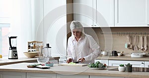 Cheerful middle aged dancing woman preparing salad in home kitchen