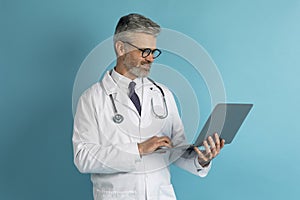 Cheerful middle aged doctor using computer on blue, consulting patient