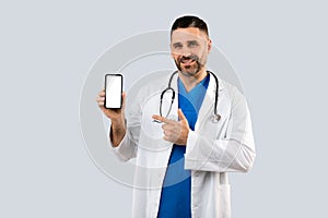 Cheerful middle aged general practitioner man in uniform pointing at cellphone blank screen, grey background, mockup