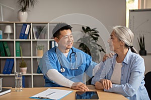 Cheerful middle aged korean man doctor supports and advises, calms old european female patient in clinic