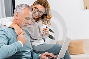 cheerful middle aged man using laptop while his smiling wife sitting near with coffee