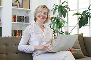 Cheerful middle aged woman using laptop while sitting on sofa at home