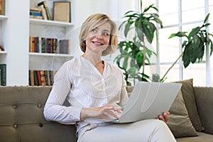 Cheerful middle aged woman using laptop while sitting on sofa at home