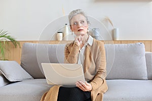 Cheerful middle aged woman using laptop while sitting on sofa at home