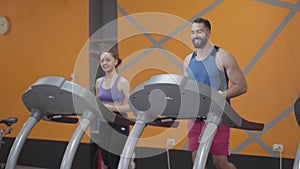 Cheerful Middle Eastern man and Caucasian woman jogging on treadmill in gym. Portrait of happy sportive multi-ethnic