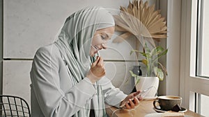 Cheerful Middle Eastern woman in hijab talking on mobile phone using earphones in cafe