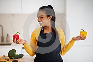 Cheerful millennial african american female in apron chooses peppers in hands at table with vegetables