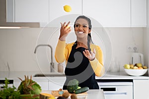Cheerful millennial african american lady in apron juggles lemon, has fun at table with organic vegetables