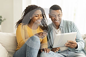 Cheerful millennial african american wife show finger at husband tablet, have video call in living room interior