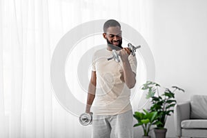 Cheerful millennial bearded african american guy in white sport wear lifts dumbbells in room interior near window
