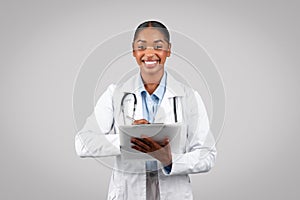Cheerful millennial black lady doctor in white coat, with stethoscope, write recipe at clipboard