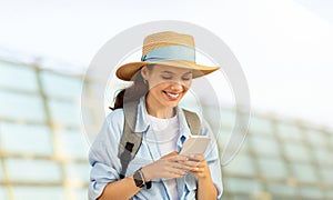 Cheerful millennial caucasian lady tourist in hat with backpack, typing on phone, use online map