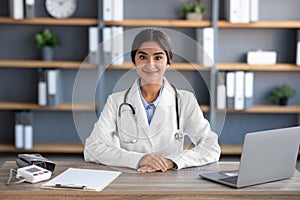 Cheerful millennial indian female doctor in white coat work at workplace in clinic office interior with laptop