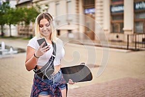 Cheerful millennial student girl walking on the streets with a skateboard in her hand and sharing a story on a smart phone while