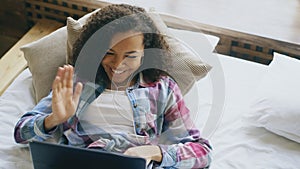 Cheerful mixed race girl having video chat with friends using laptop camera while lying on bed