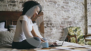 Cheerful mixed race girl typing on laptop computer for sharing social media sitting in bed at home