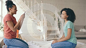 Cheerful mixed race woman photographing his girlfriend using smartphone camera sitting on bed in bedroom at home