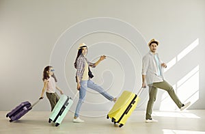 Cheerful mom, dad and their daughter have fun walking with suitcases along the gray wall.