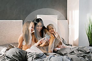 Cheerful mom and daughters are laughing using a smartphone app, at home, sitting on the bed