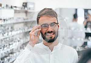 Cheerful mood. Stylish man with beard choosing glasses in the optics store