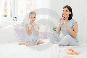 Cheerful mother and daughter dressed in pyjamas, have breakfast in morning, drink milk shake with doughnuts, sit crossed legs on c