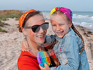 Cheerful mother and daughter smiling walking on sea beach. Happy family. Cute little child playing rainbow pop it toy