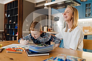 Cheerful mother helping her little son with homework in cozy kitchen at home