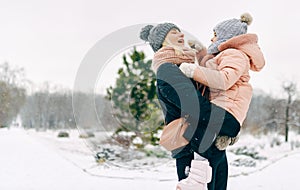 Cheerful mother holding her child in arms, smiling broadly during walking in the park on snowing day. Young woman and her daughter photo
