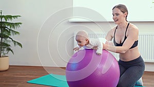Cheerful mother playing with infant, making baby fitness exercises on big ball