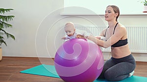 Cheerful mother playing with infant, making baby fitness exercises on big ball