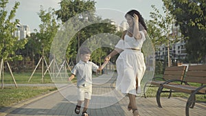 Cheerful mother and son running along sunny alley in summer park. Wide shot portrait of happy Middle Eastern woman and
