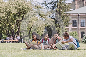 Cheerful multicultural schoolkids sitting on lawn and reading books