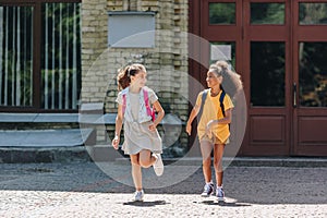 Cheerful multiethnic schoolgirls smiling while running in schoolyard