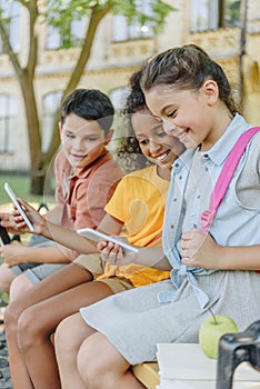Cheerful multiethnic schoolkids using smartphones while sitting on bench in schoolyard