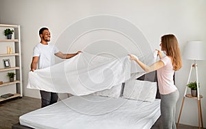 Cheerful multiracial couple making bed in morning, having fun at home
