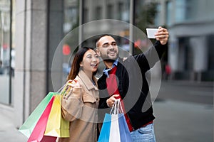 Cheerful multiracial couple with shopper bags taking selfie on smartphone in front of big supermarket