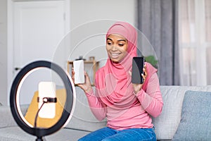 Cheerful muslim black blogger filming video review of two new smartphones, talking to cellphone webcamera on ring lamp