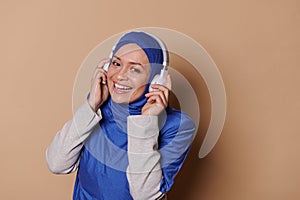 Cheerful Muslim woman in blue hijab, listening a playlist on headphones and smiling at camera over beige background